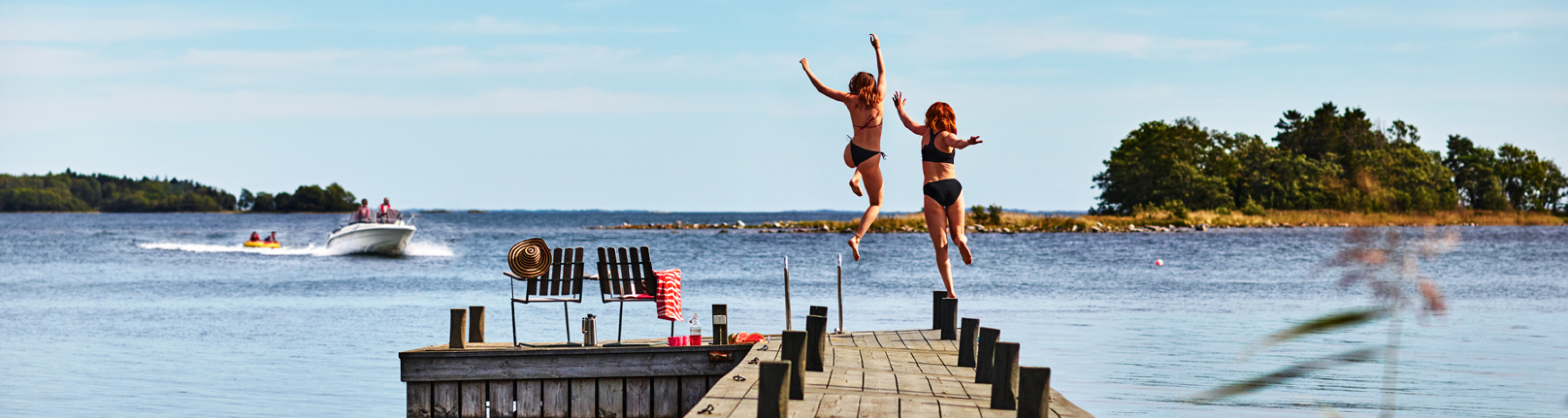 Allt för sjön Båtmässan - en bild på en badbrygga med två personer som hoppar i vattnet. En motorbåt och en udde syns även i bild. 